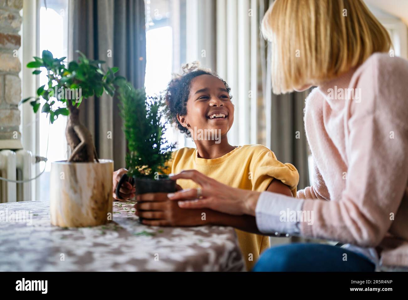 Fröhliche weiße Mutter und ihre adoptierte afroamerikanische Tochter, die sich um Pflanzen im Haus kümmert. Stockfoto