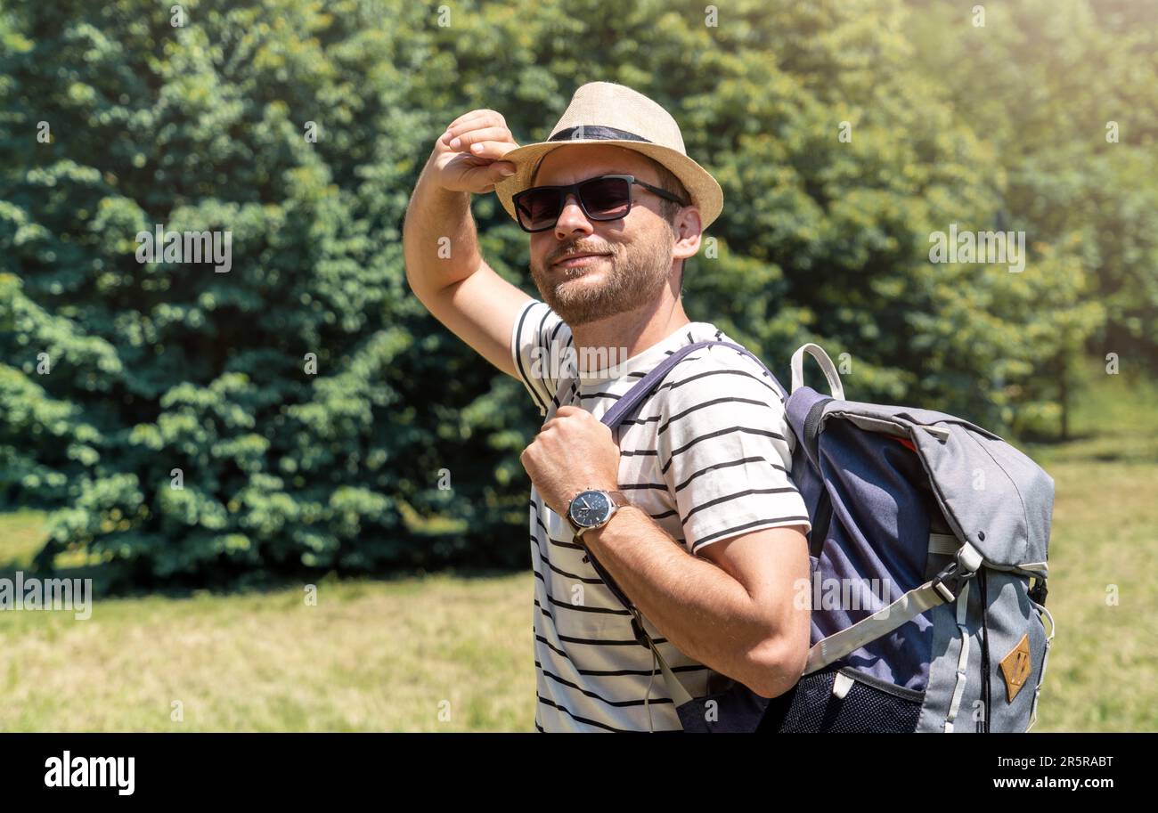 Erwachsener Mann mit Sonnenbrille, Hut und Rucksack, der den Sommer im Stadtpark genießt, in die Kamera schaut und lächelt. Stockfoto