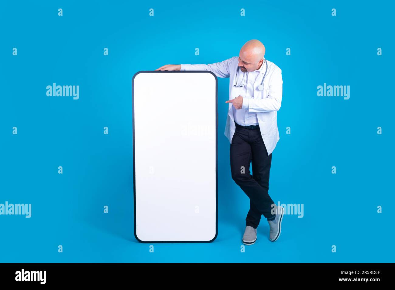 Männlicher Arzt mittleren Alters, lehnt sich und zeigt auf ein großes, riesiges Handy mit leerem Bildschirm. Arzt in voller Größe, der auf isoliertem blauem Hintergrund steht. Stockfoto
