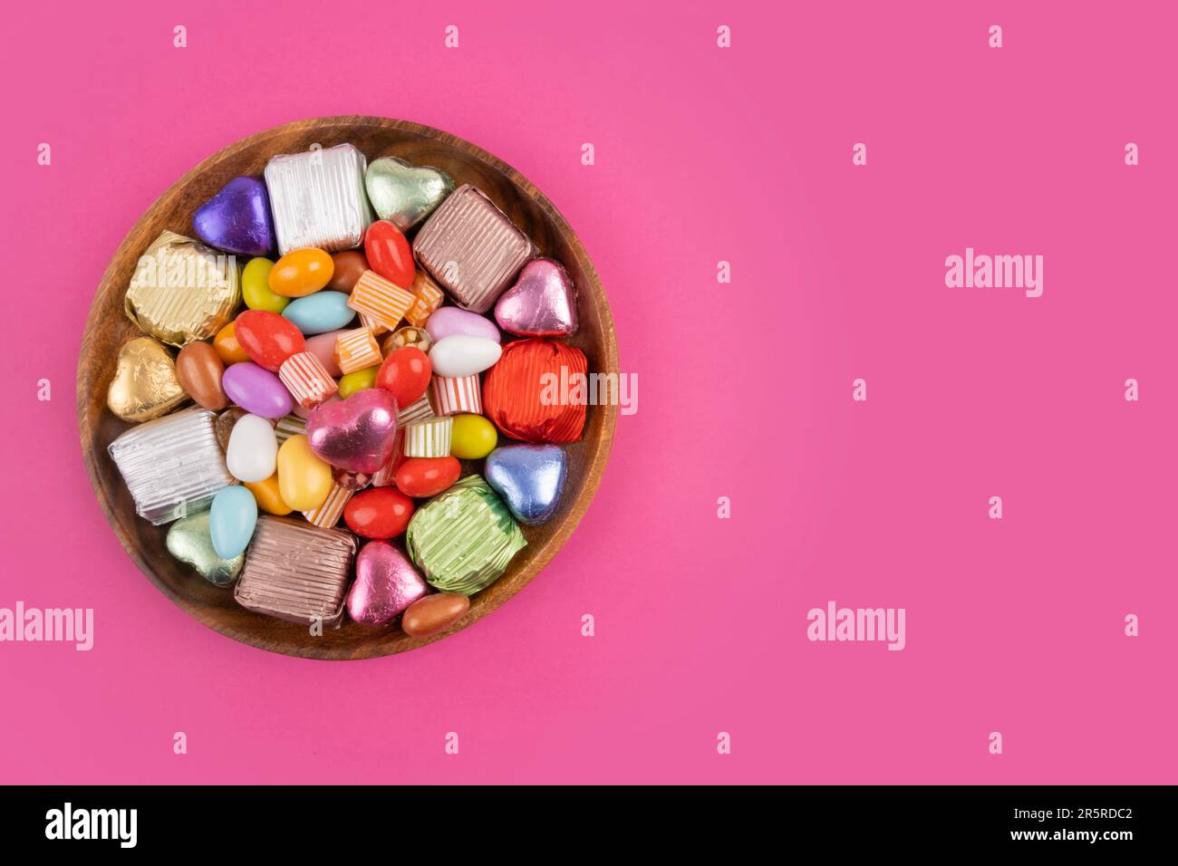 Foto von oben mit einer Schüssel Süßigkeiten. Bunte Süßigkeiten auf dem Holzteller. Isolierter rosafarbener Hintergrund, Kopierraum für Begrüßungen. Luxuriöse Schokolade in Geschenkverpackung. Stockfoto