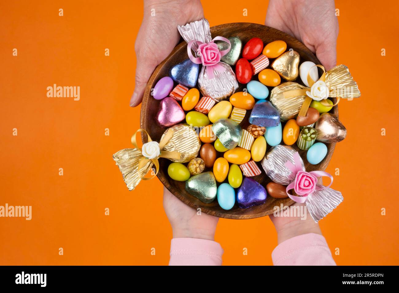 Die Hände von Frau und Kind halten eine Schüssel süß. Bild von oben: Umwickelte Luxus-Schokolade und Mandeldragee in Holzschale. Isolierter orangefarbener Hintergrund. Stockfoto