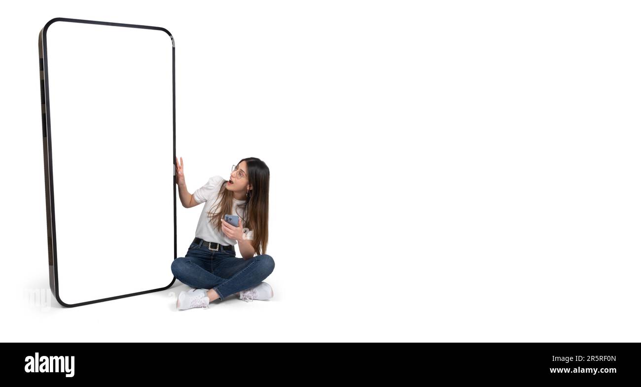 Aufgeregte Frau, die in der Nähe eines großen Smartphones mit leerem Bildschirm sitzt. Ganzkörperjunge kaukasische Mädchen, die ein großes Handy sieht und sich berührt. Mobile App. Stockfoto