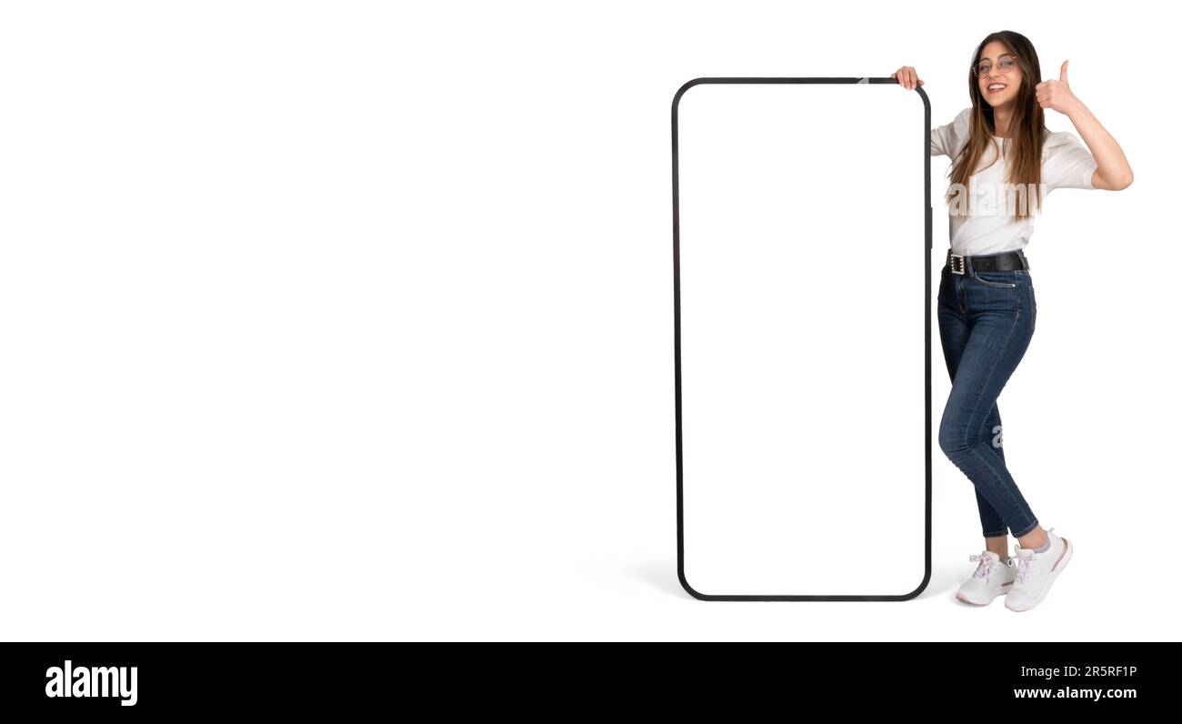 Ganzkörperfoto einer Frau, die eine mobile Anwendung oder Website empfiehlt. Kaukasische Frau, die großes Smartphone mit leerem, leerem Bildschirm-Mockup anlehnt. Daumen hoch. Stockfoto