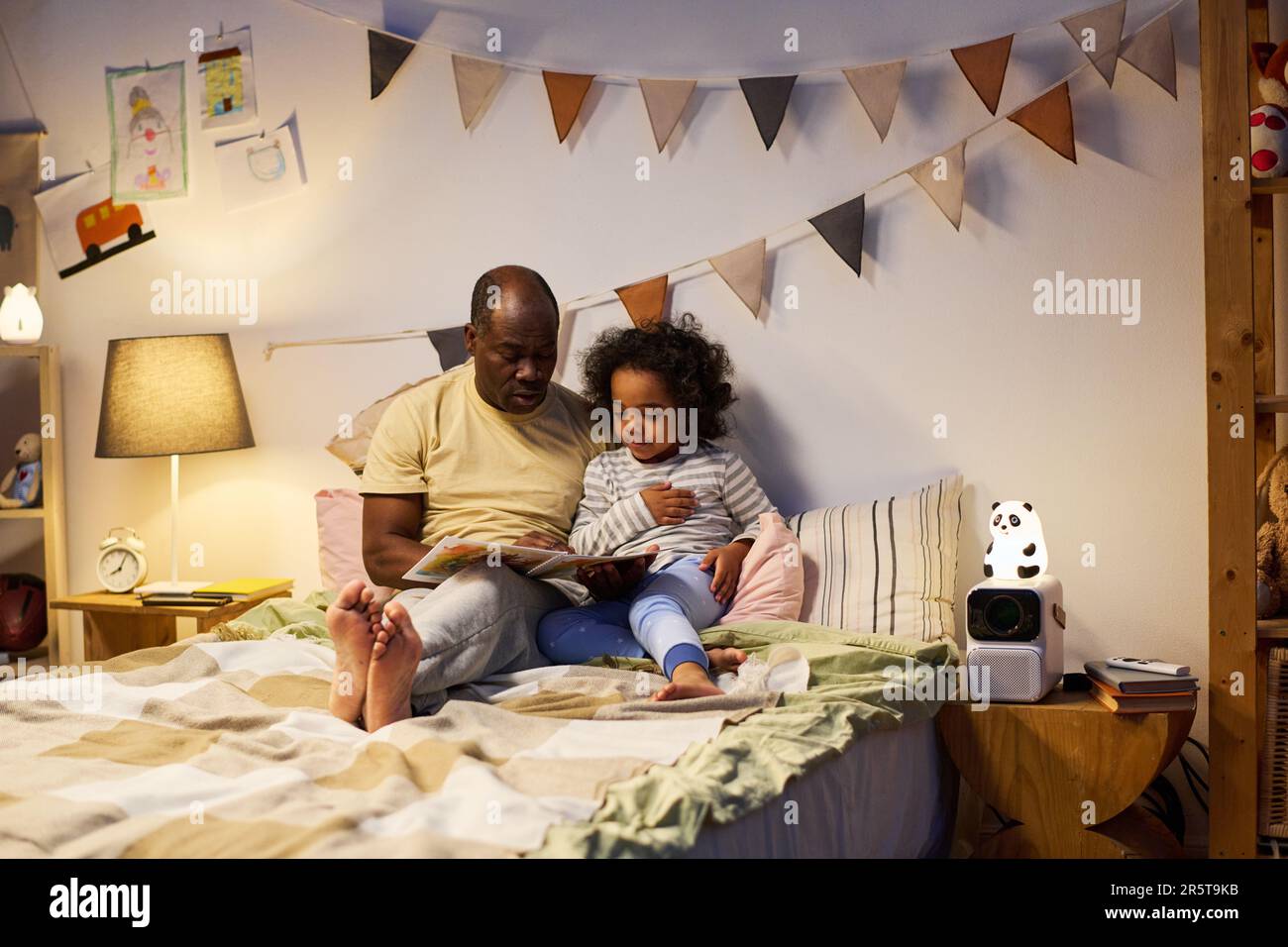 Ein afroamerikanischer Vater liest seinem Sohn vor dem Schlafen ein Buch, während sie im Bett im Schlafzimmer sitzen Stockfoto
