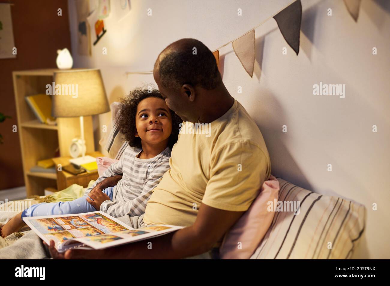 Ein afroamerikanischer Vater, der vor dem Schlafen seinem Kind Märchen vorliest, sie schauen sich Bilder an und reden miteinander Stockfoto