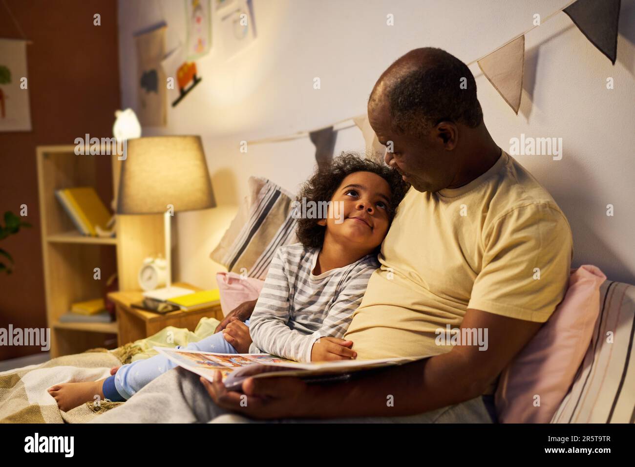 Ein afroamerikanischer Vater liest dem Kind vor dem Schlafen ein Buch vor, während er ihn im Bett umarmt Stockfoto