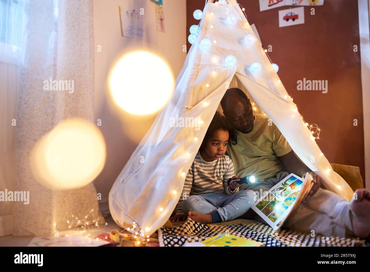 Dad liest seinem Kind ein Buch vor, während es in einem handgemachten Zelt im Zimmer sitzt Stockfoto