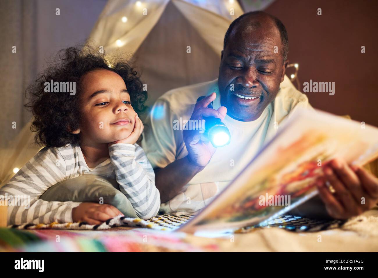 Ein afroamerikanischer Vater, der Bücher und Taschenlampen in der Hand hält und seinem kleinen Sohn Geschichten erzählt, während sie im Zimmer auf dem Boden liegen Stockfoto