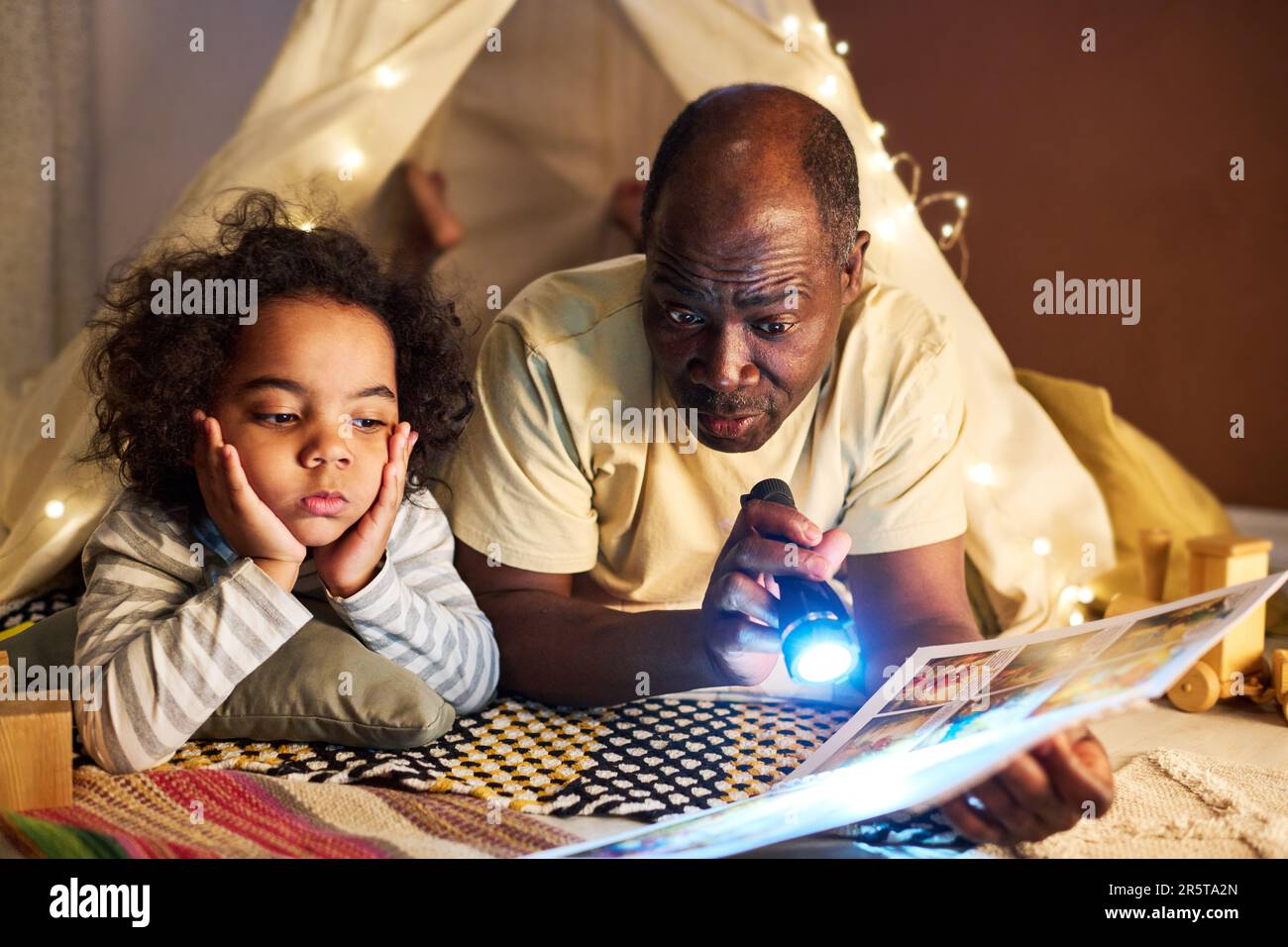 Ein afroamerikanischer Vater liest seinem Sohn mit Taschenlampe ein Buch, während sie auf dem Boden in einem hausgemachten Zelt liegen Stockfoto