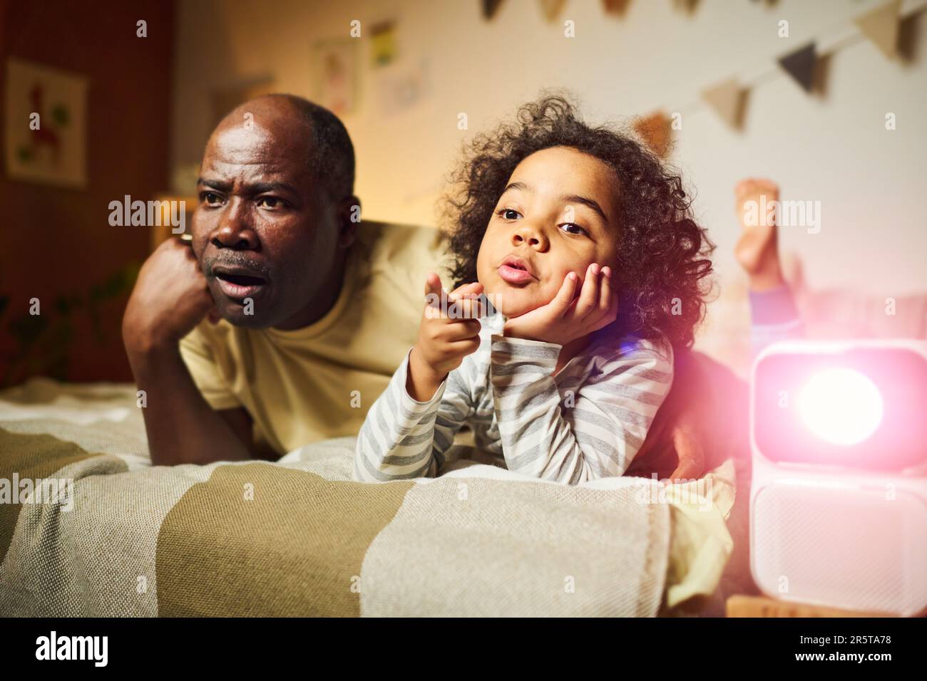 Ein afroamerikanischer Vater, der seinem Sohn einen Film auf dem Projektor zeigt, während sie im Bett im Schlafzimmer liegen Stockfoto