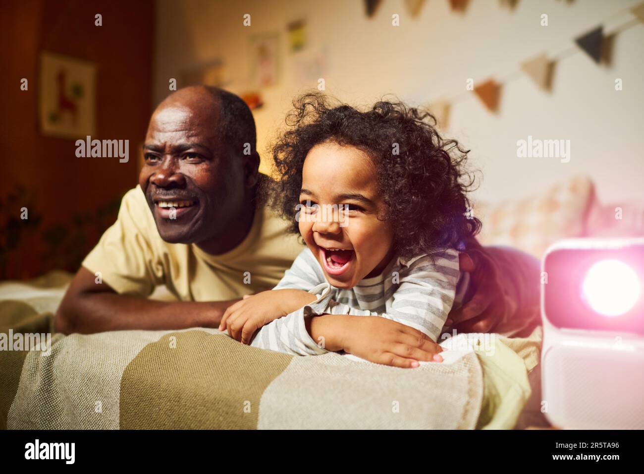 Afroamerikanische Familie mit zwei Personen, die sich zusammen lustige Filme auf dem Projektor ansehen, während sie im Zimmer auf dem Bett lag Stockfoto