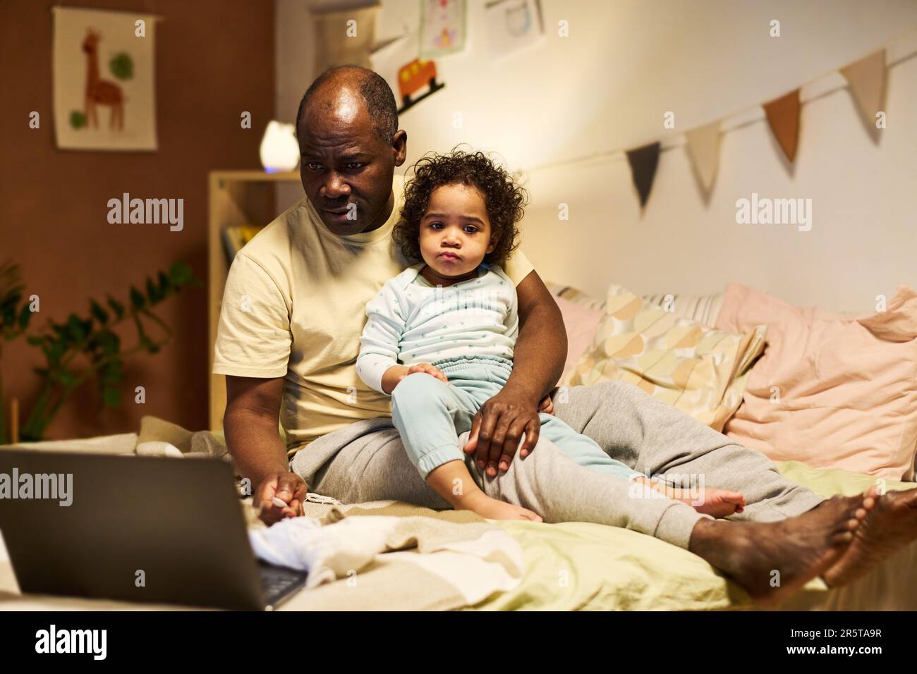 Afroamerikanischer Vater, der online auf einem Laptop arbeitet, während er mit dem Baby zu Hause sitzt Stockfoto