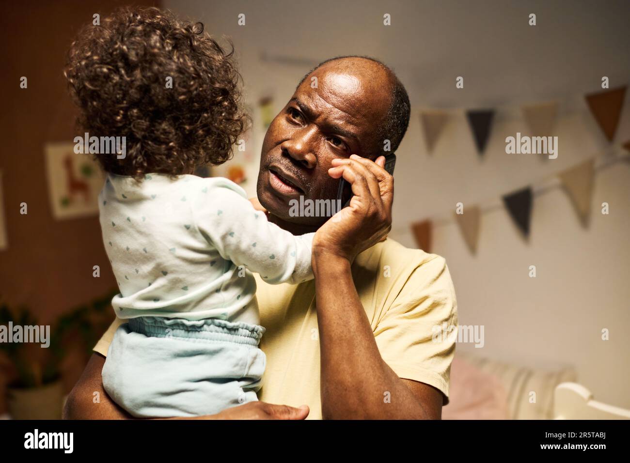 Ein seriöser afroamerikanischer Vater, der zu Hause mit seinem Baby am Telefon sitzt Stockfoto