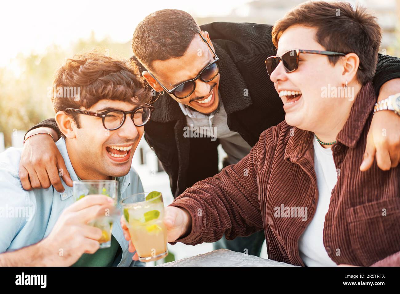 Ein afroamerikanischer Mann, eine nicht binäre Frau und eine Kaukasierin treffen sich in einer Bar im Freien, genießen Mojito-Cocktails und teilen Lachen. Stockfoto