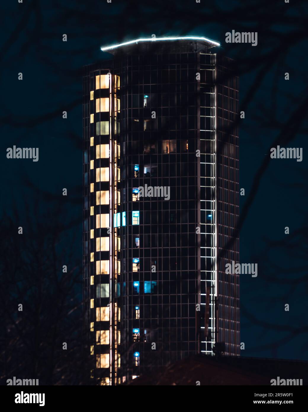 Ein Hochhaus, beleuchtet in einem lebendigen blauen Licht, teilweise von Bäumen in einer nächtlichen Umgebung verdeckt Stockfoto