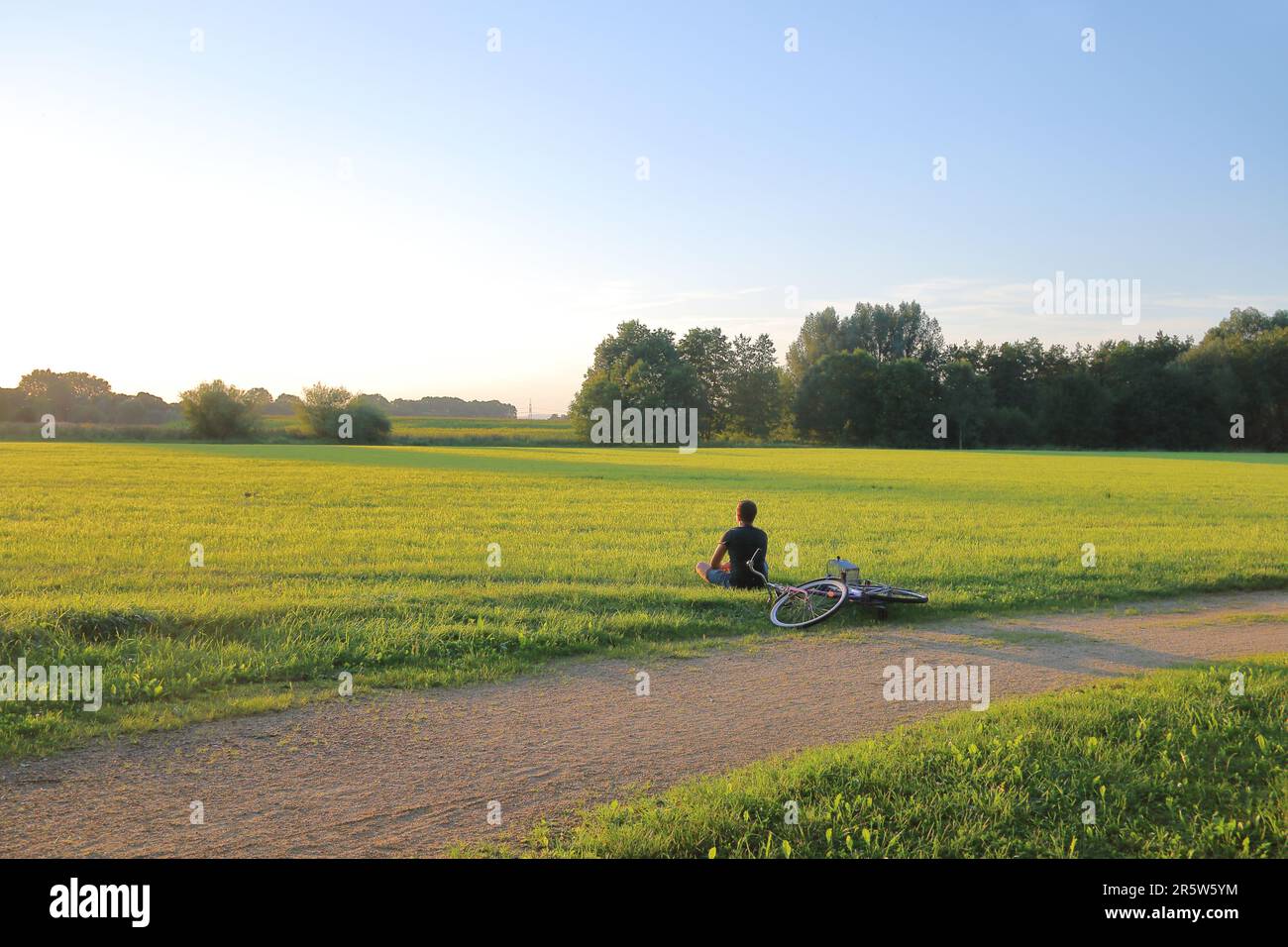 Auf dem Foto sah man einen jungen Mann, der auf der Wiese saß und den Sonnenuntergang bewunderte. In der Nähe des sichtbaren Fahrrads, das auf dem Gras liegt. Stockfoto