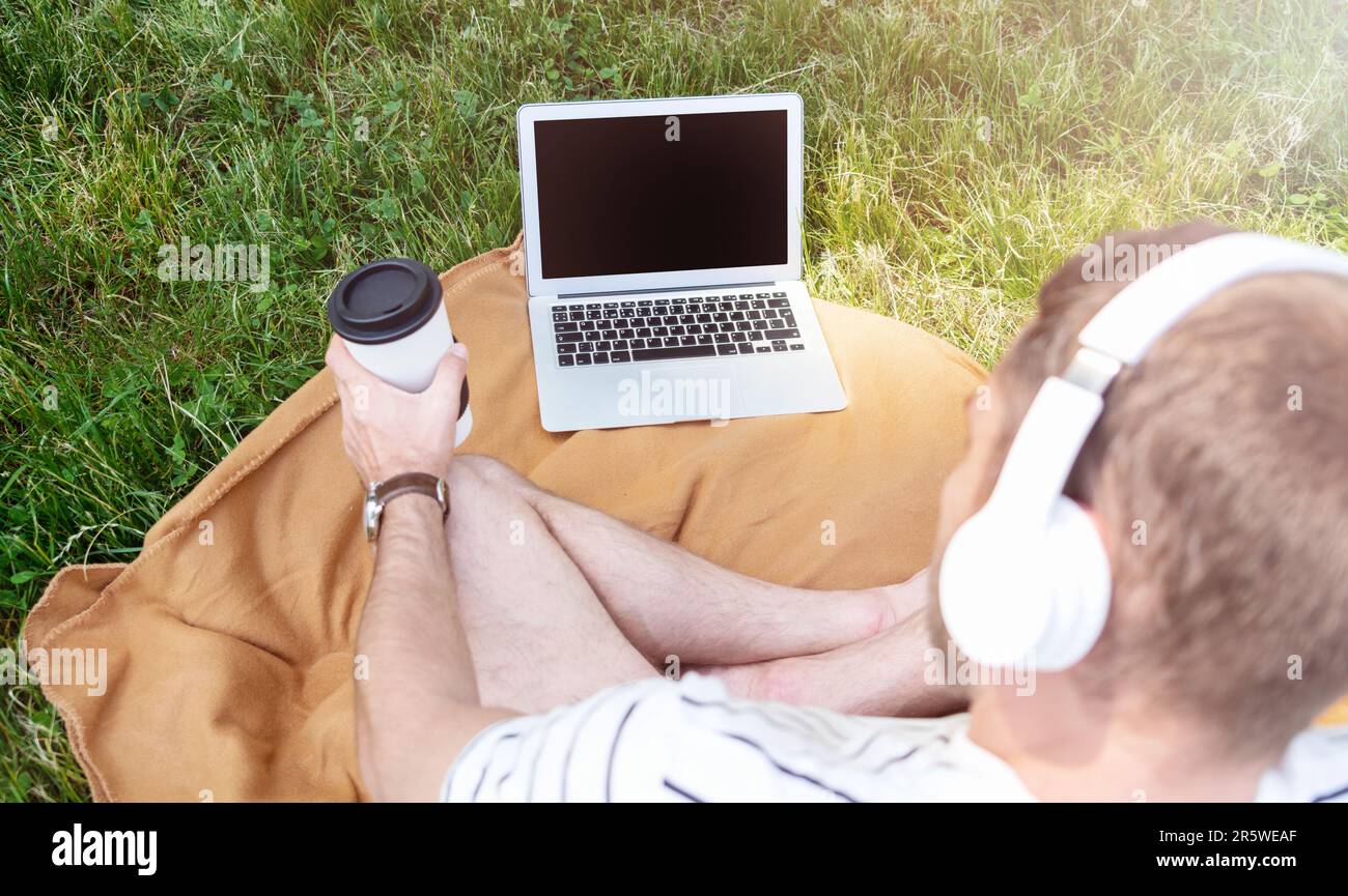 Mann mit Kopfhörern, der sich entspannt auf dem Rasen im Stadtpark sitzt und sein Laptop nutzt, Wochenend-Freizeitaktivitäten Stockfoto