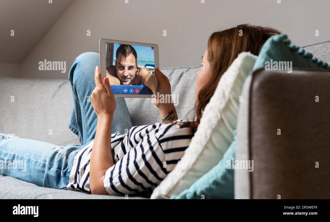 Junge Frau, die zu Hause auf dem Sofa liegt und ihren Freund per Video mit einem digitalen Tablet anruft. Stockfoto