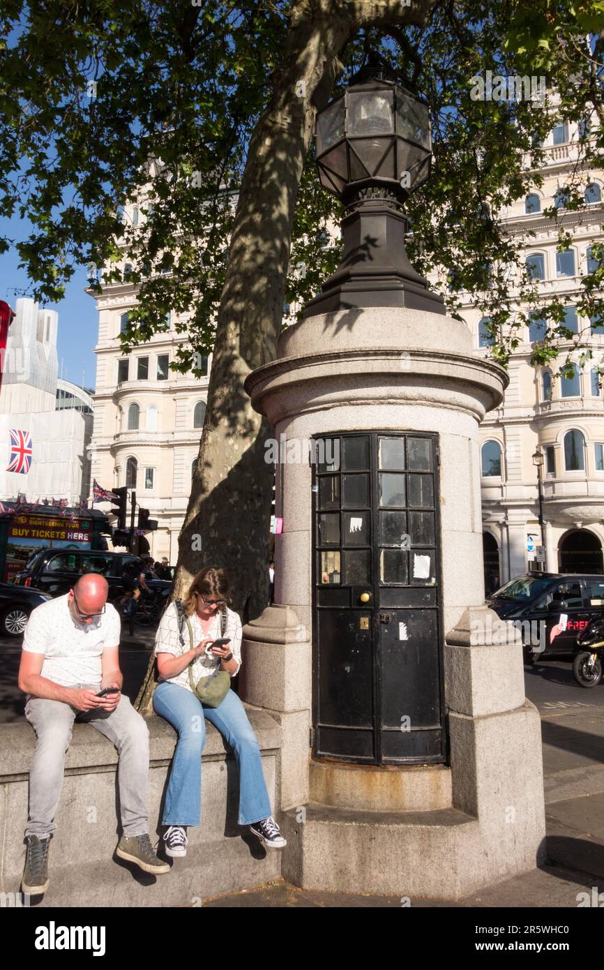 Touristen auf ihren Mobiltelefonen, die an einem heißen Sommertag neben der kleinsten Polizeistation Londons sitzen, Trafalgar Square, London, WC2, England, UK Stockfoto