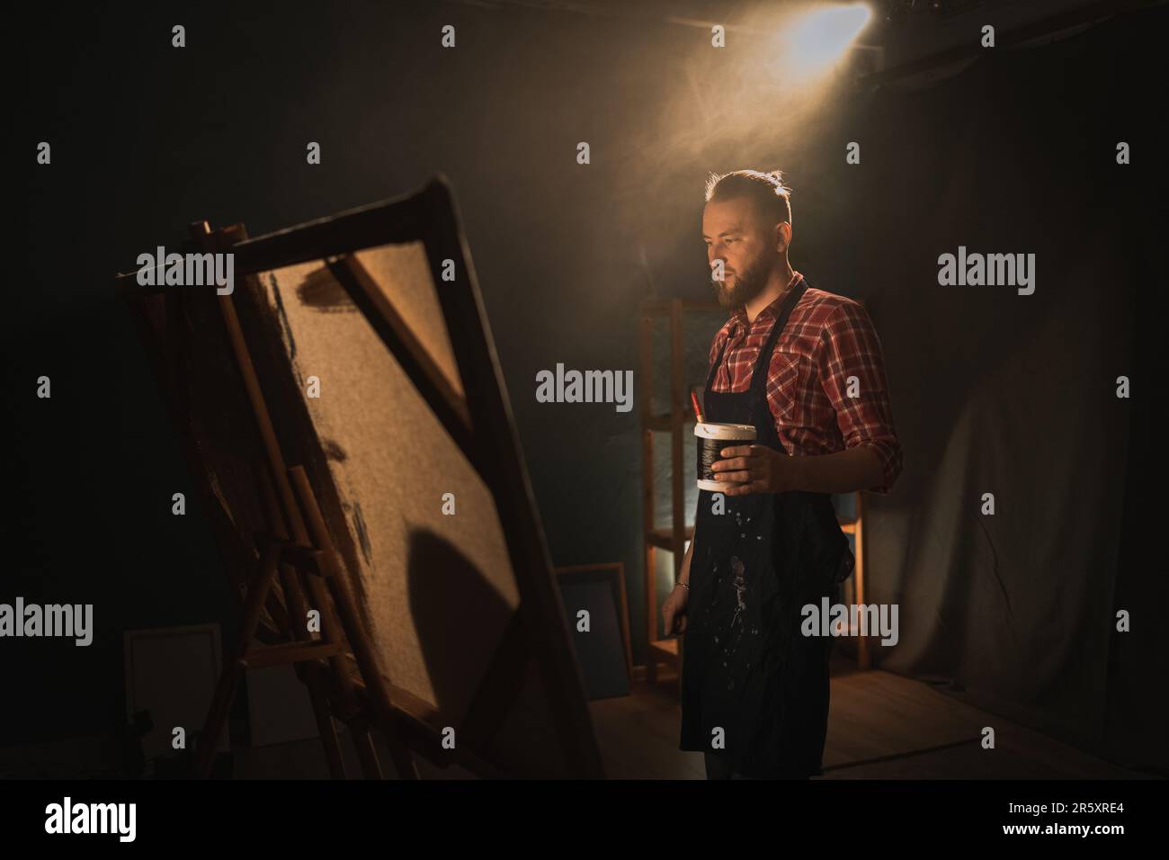 Talentierter männlicher Künstler arbeitet an abstrakten Ölgemälden und kreiert ein modernes Meisterwerk in einem dunklen Kreativstudio mit großer Leinwand. Stockfoto