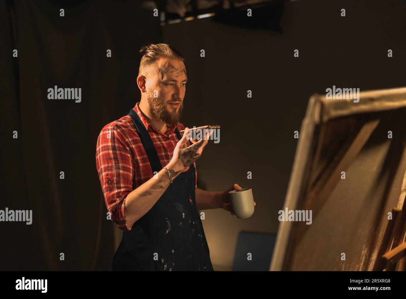 Männlicher Künstler steht in seinem modernen kreativen Studio mit großer Leinwand, hält Telefongespräche mit einem Freund über Lautsprecher, macht Spracherkennung oder Stockfoto