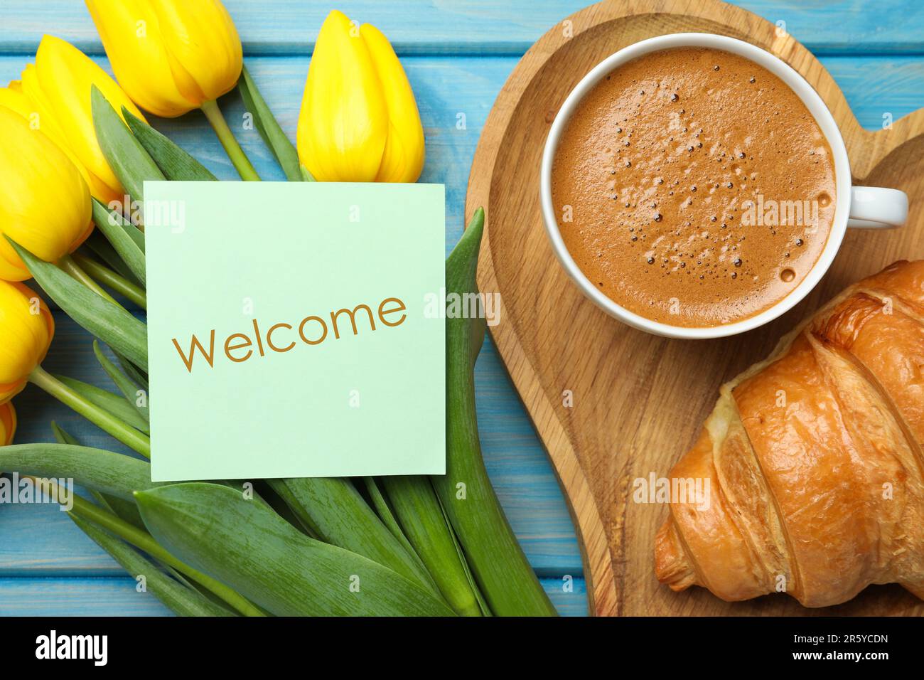 Begrüßungskarte, wunderschöne gelbe Tulpen, Croissant und eine Tasse aromatischen Kaffee auf einem hellblauen Holztisch, flach liegend Stockfoto