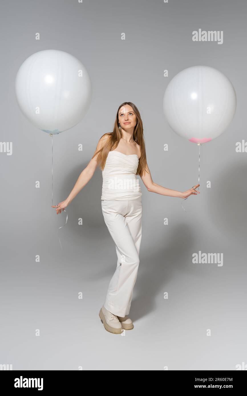 Eine trendige und schwangere Frau blickt während der Feier weg, während sie weiße festliche Ballons hält, und das Geschlecht zeigt eine Überraschungsparty auf Grau Stockfoto