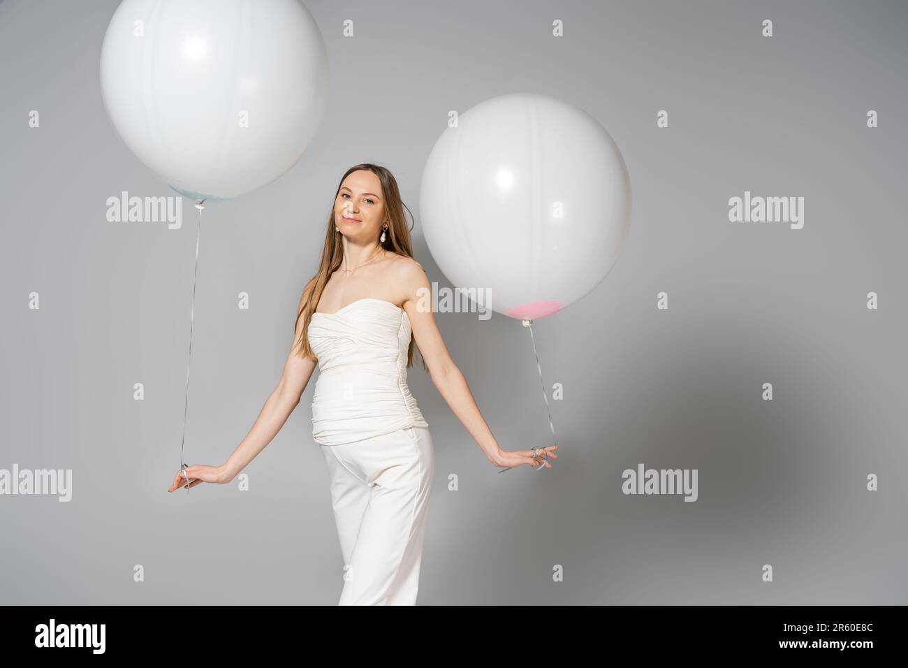 Eine positive und modische schwangere Frau, die in die Kamera schaut, während sie weiße festliche Ballons hält, während das Geschlecht eine Überraschungsparty auf grauem Hintergrund enthüllt, Stockfoto