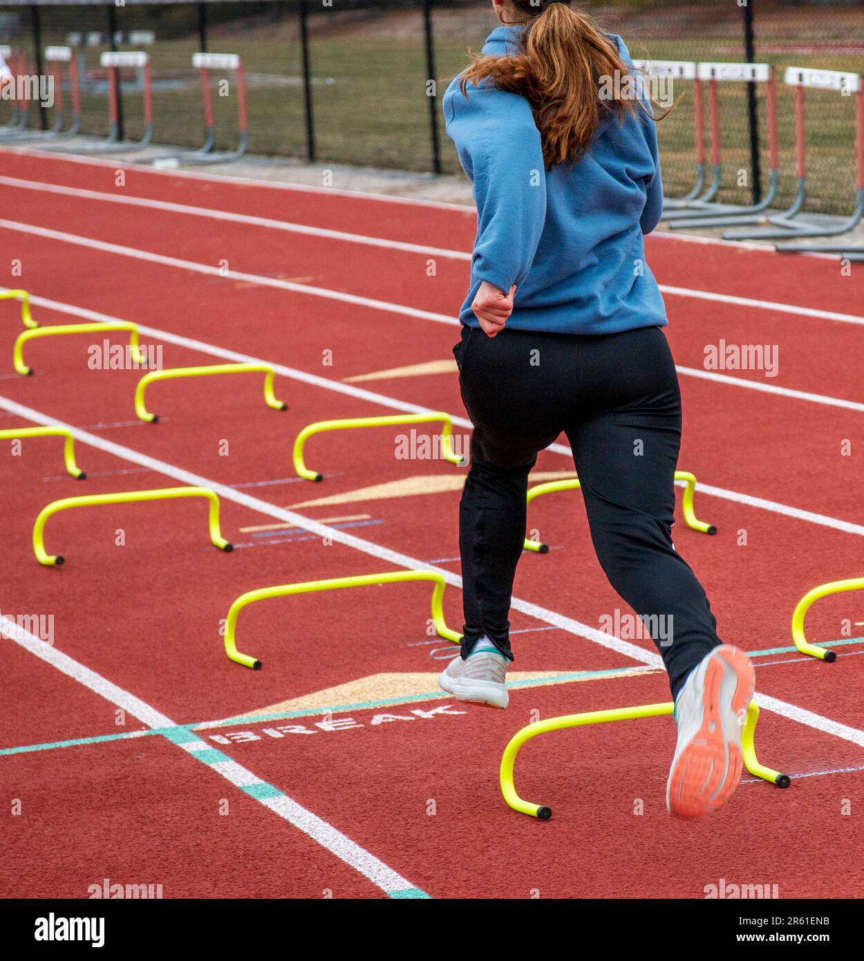 Rückansicht eines Highschool-Leichtathletikmädchens, das während des Trainings für Geschwindigkeit und Beweglichkeit auf einer roten Strecke über kleine gelbe Hürden auf der Spur springt. Stockfoto