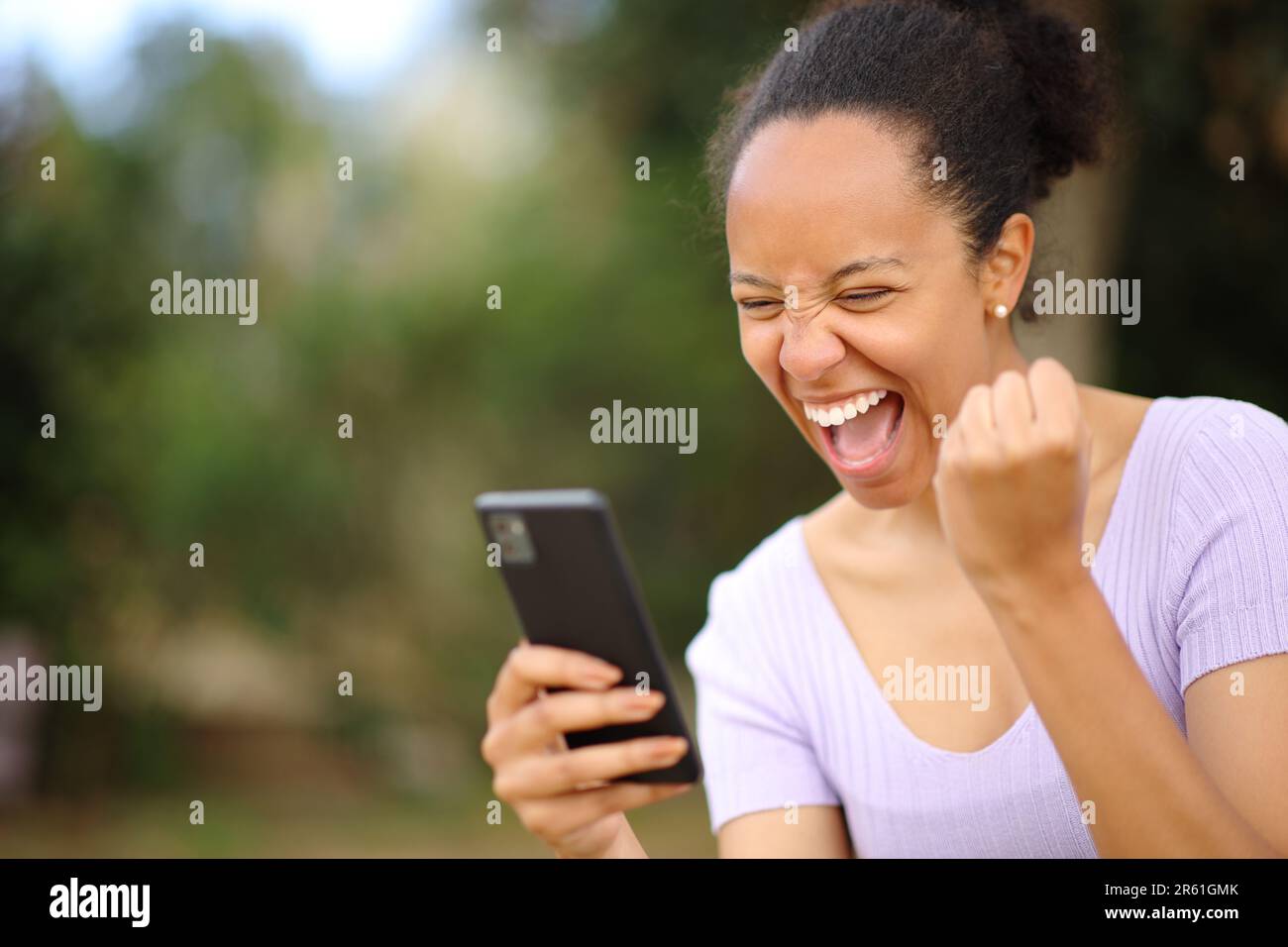 Aufgeregte schwarze Frau feiert gute Nachrichten am Telefon in einem Park Stockfoto
