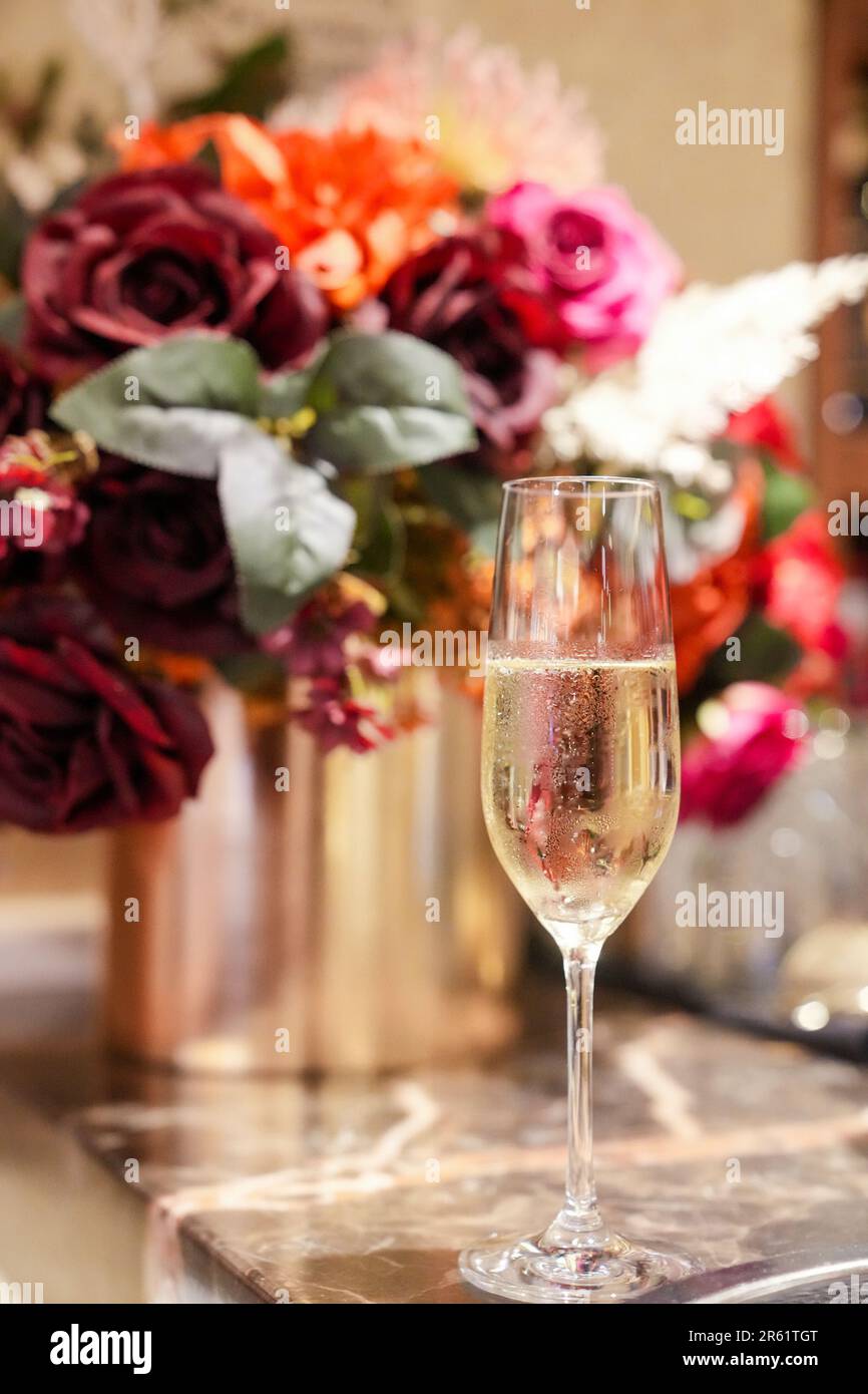 Ein kristallklares Glas Sekt auf einem Tisch, umgeben von einem Strauß farbenfroher Blumen in einer Glasvase Stockfotografie
