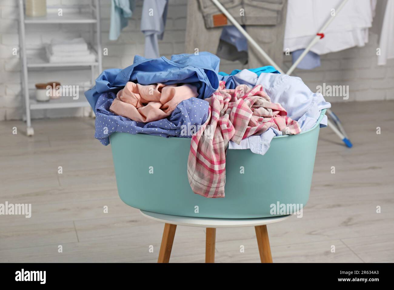 Wäschekorb aus Plastik, überfüllt mit Kleidung auf weißem Hocker im Innenbereich Stockfoto