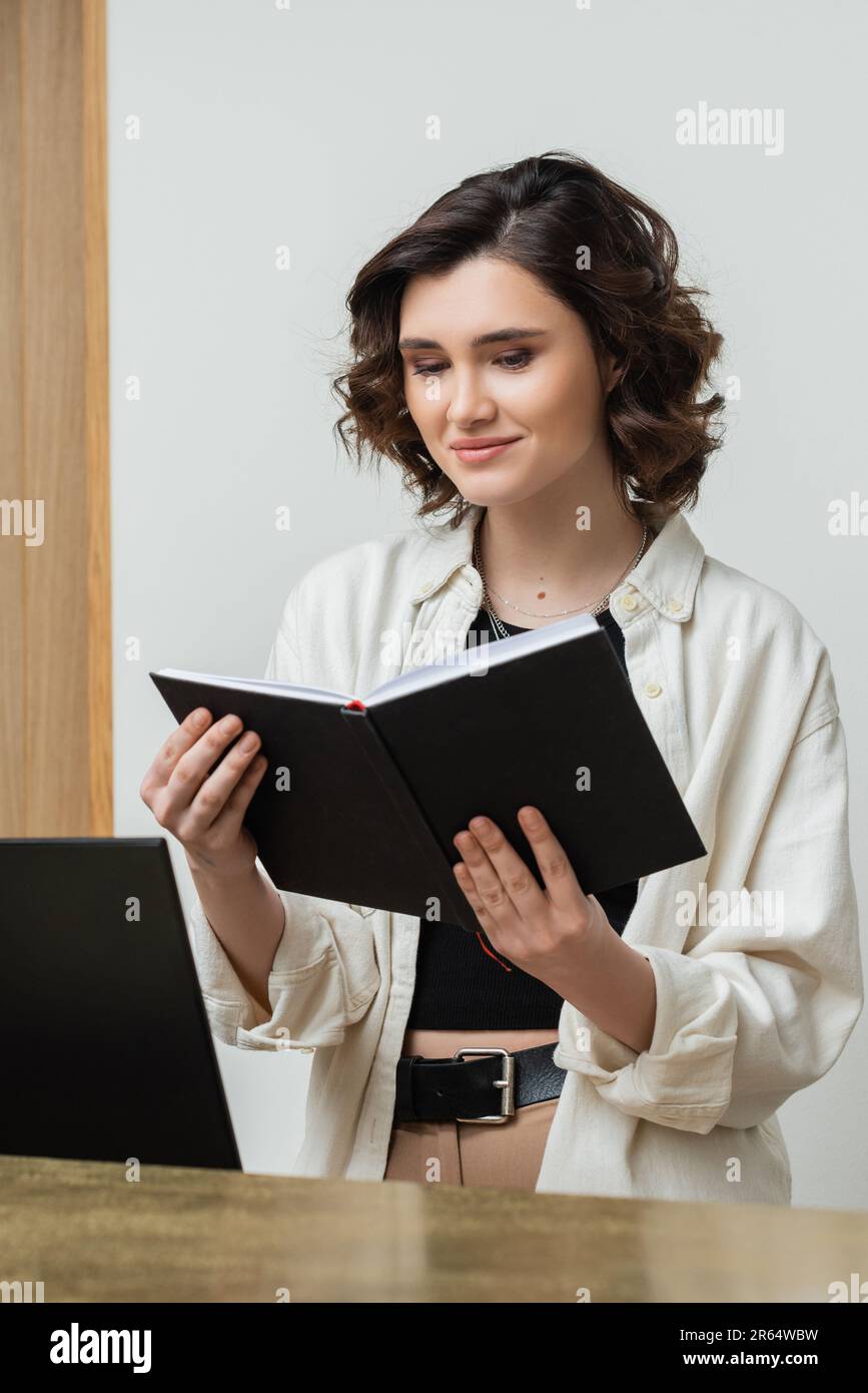 Fröhlicher Concierge in trendigen, lässigen Kleidern und mit welligen braunen Haaren, der in der Nähe des Computerbildschirms auf das Notebook blickte, während er an der Rezeption arbeitete Stockfoto