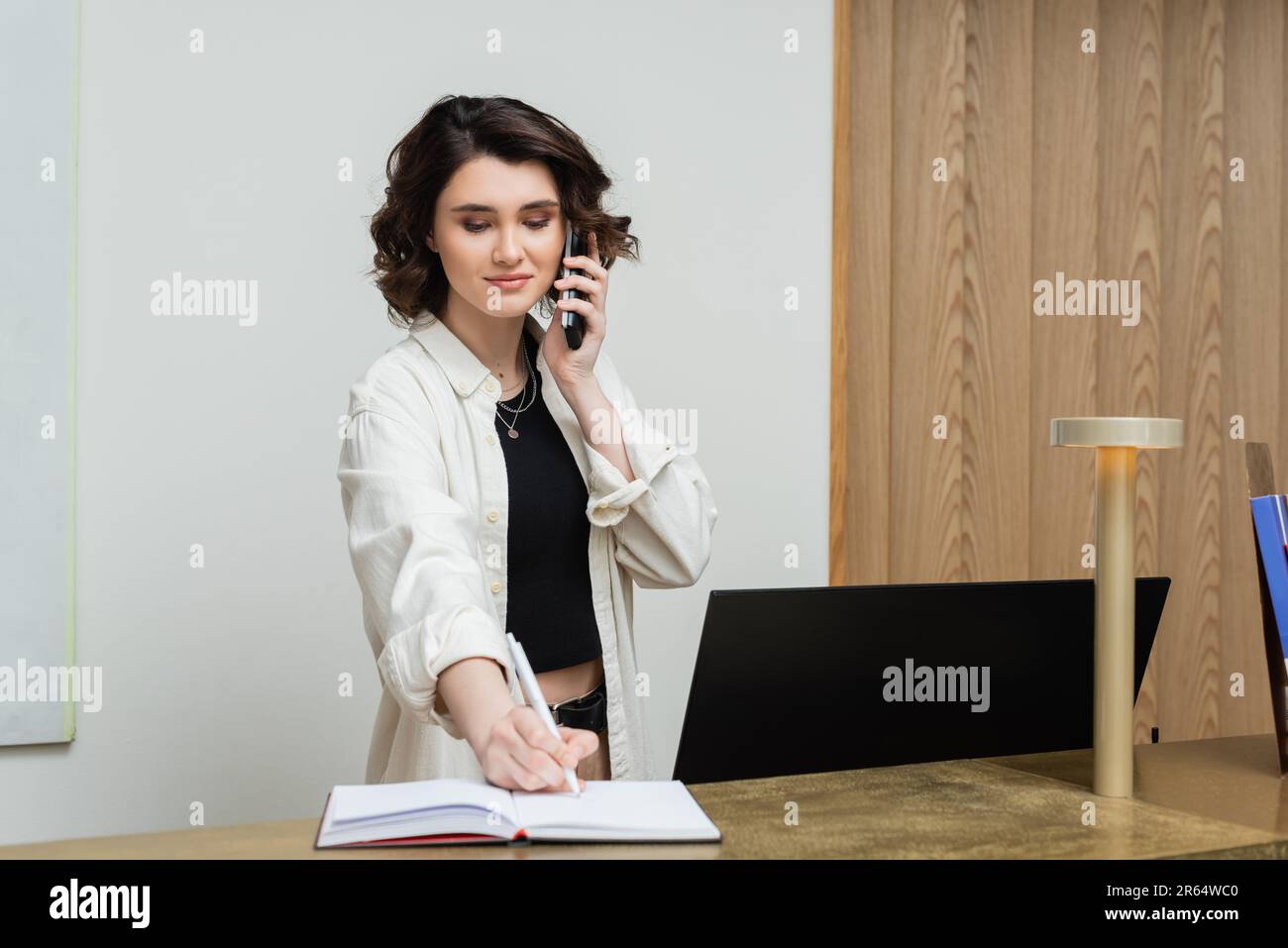 Freudiger Rezeptionist mit welligen braunen Haaren, der an der Rezeption arbeitete, in einem Notizbuch schrieb und am Telefon in der Nähe des Computerbildschirms in der Lobby sprach Stockfoto