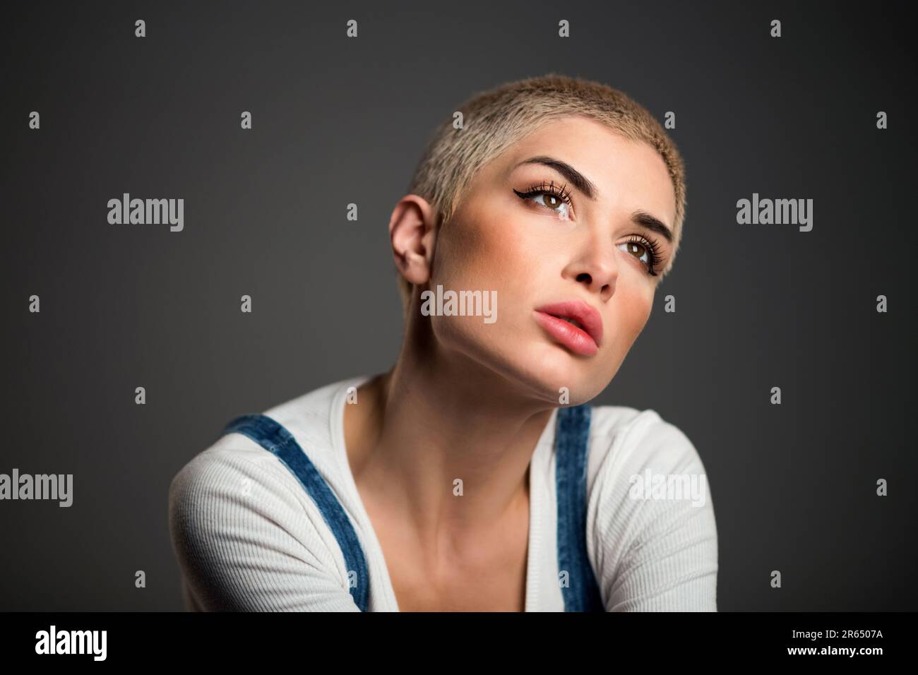 Das nachdenkliche junge, stilvolle weibliche Modell mit kurzen blonden Haaren und legerer Kleidung blickt traumhaft vor schwarzem Hintergrund weg Stockfoto