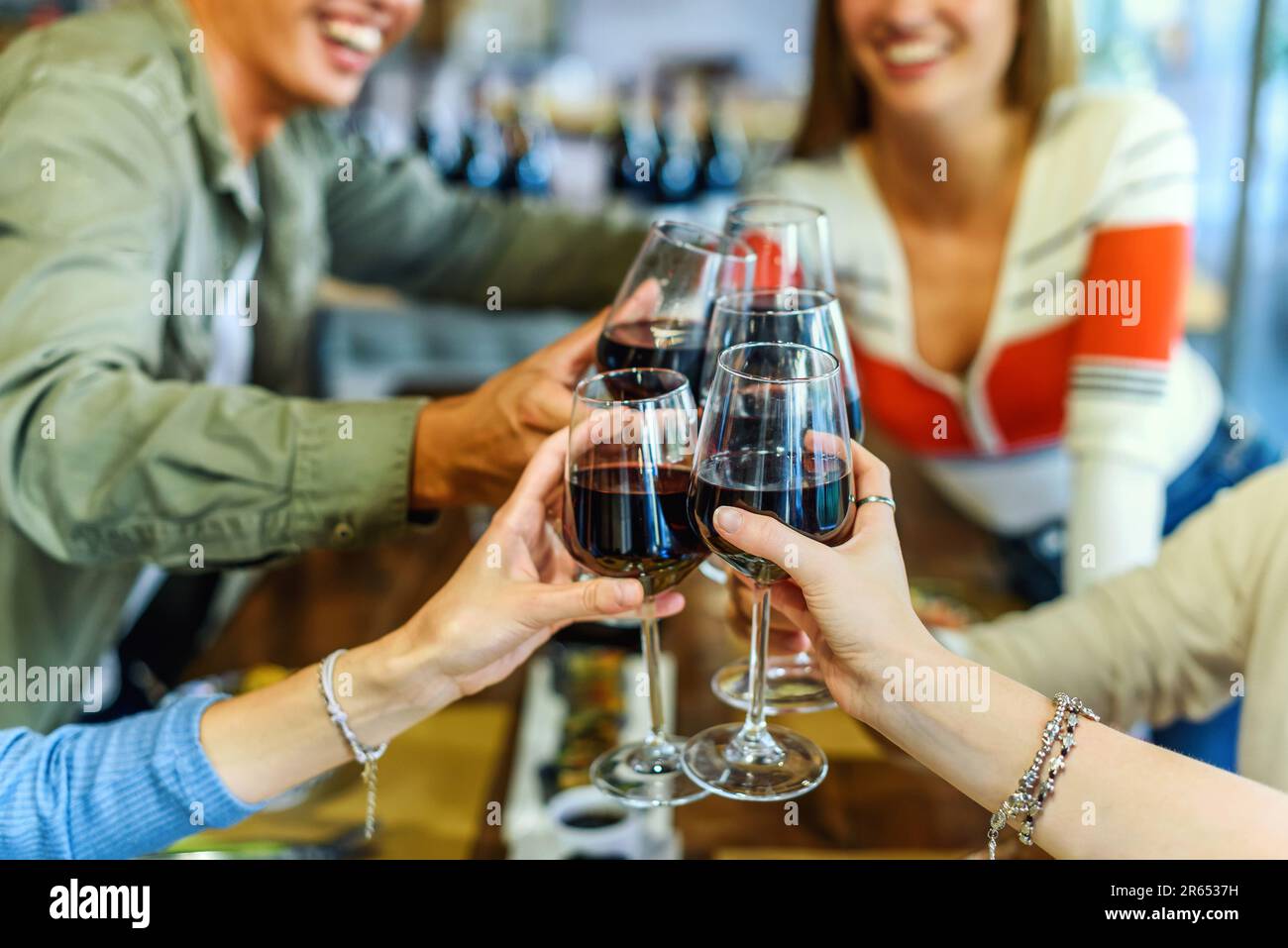 Ernten Sie und nicht erkennbare, fröhliche Freunde in lässiger Kleidung, sitzen Sie zusammen am Tisch und klappern Sie mit Gläsern Rotwein, während Sie die Zeit verschwommen verbringen Stockfoto