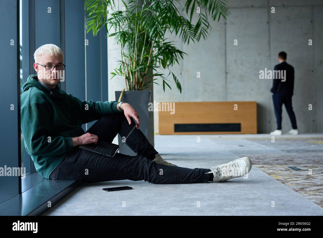 Ein junger blonder Mann in einem modernen Büro sitzt am Fenster, vertieft in seine Arbeit auf einem Laptop Stockfoto