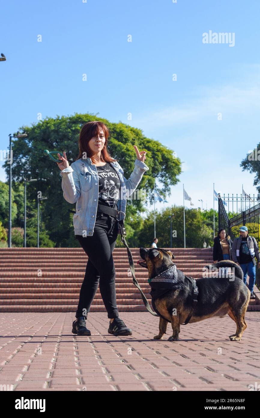 Eine junge lateinisch-kolumbianische Frau, die mit ihrem Hund im Park in die Kamera guckt und mit gekreuzten Händen und Beinen Frieden und Liebe signiert Stockfoto