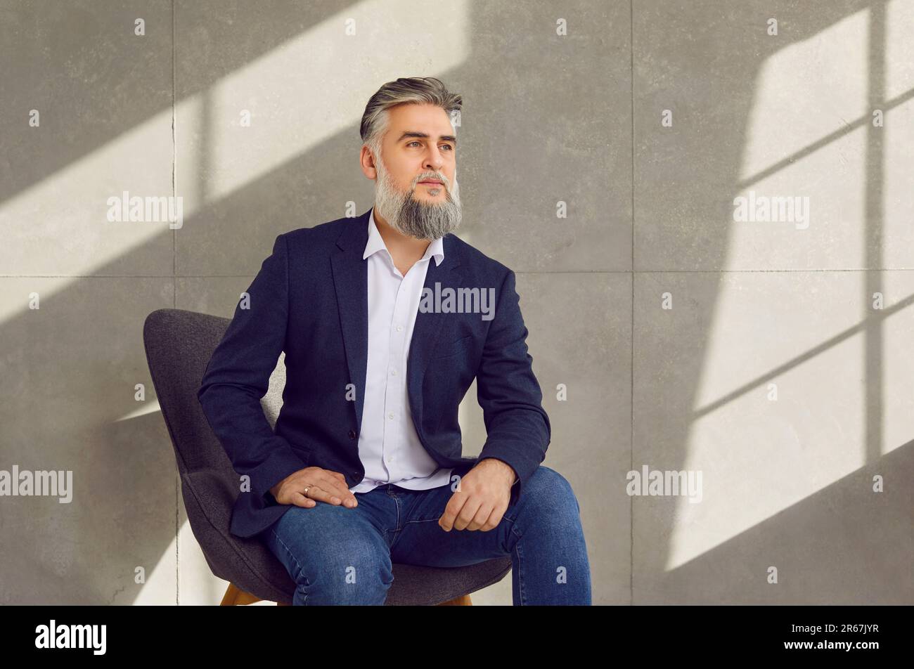 Selbstbewusster, bärtiger, reifer Geschäftsmann, der auf einem Stuhl sitzt, in leerem Raum auf grauem Hintergrund. Stockfoto