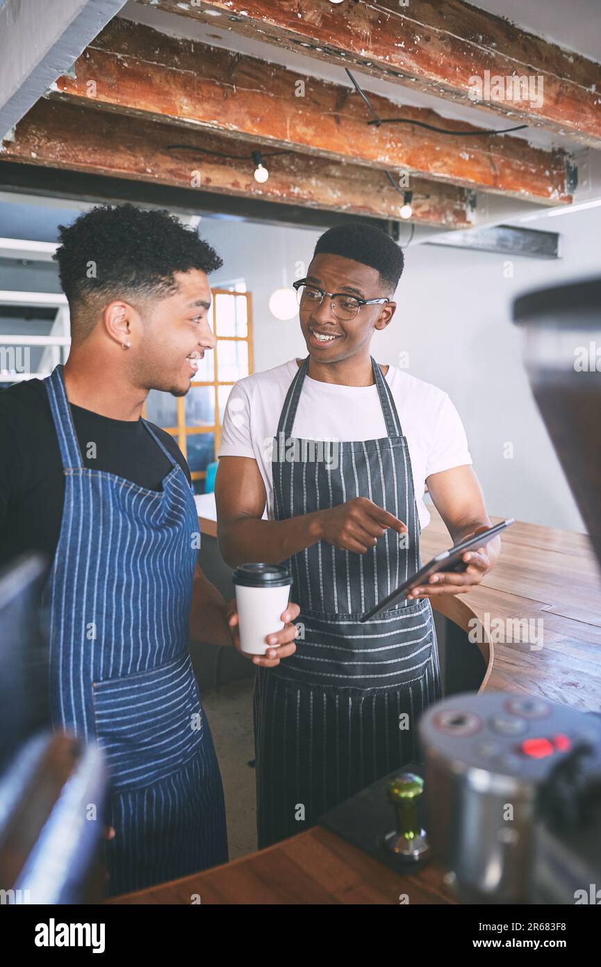 Bestellungen mit guter alter Teamarbeit zu bekommen. Zwei junge Männer, die ein digitales Tablet verwenden, während sie Kaffee in einem Café zubereiten. Stockfoto