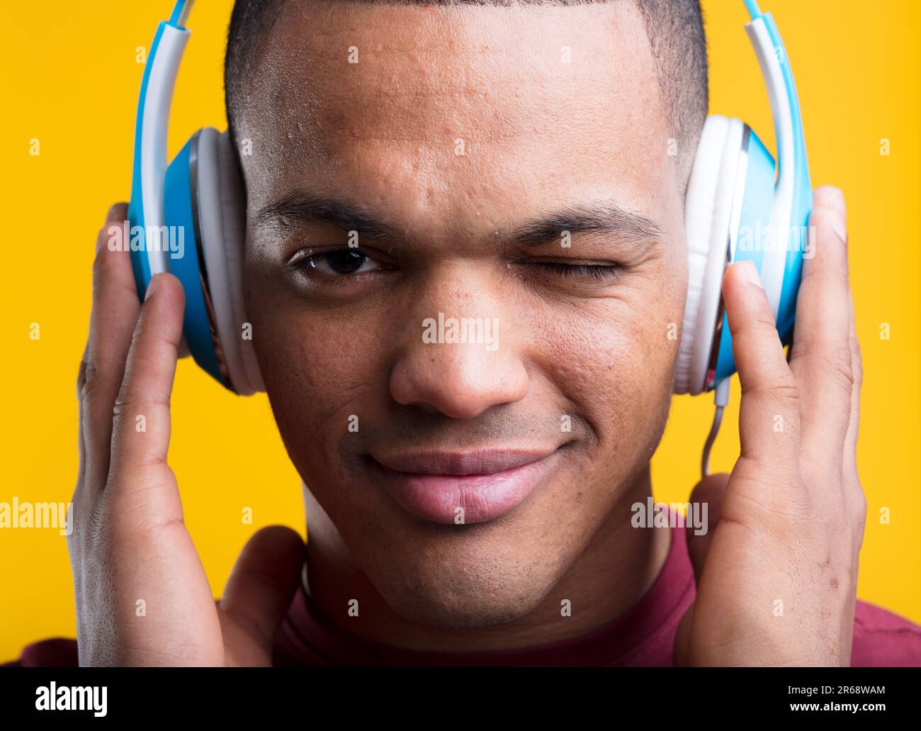 Der junge Mann zwinkert, während er leidenschaftlich Musik oder Podcasts hört. Er hält blaue Kopfhörer auf gelbem Hintergrund in der Hand. Kurze rasierte Haare, dunkelbraune Haut, b Stockfoto