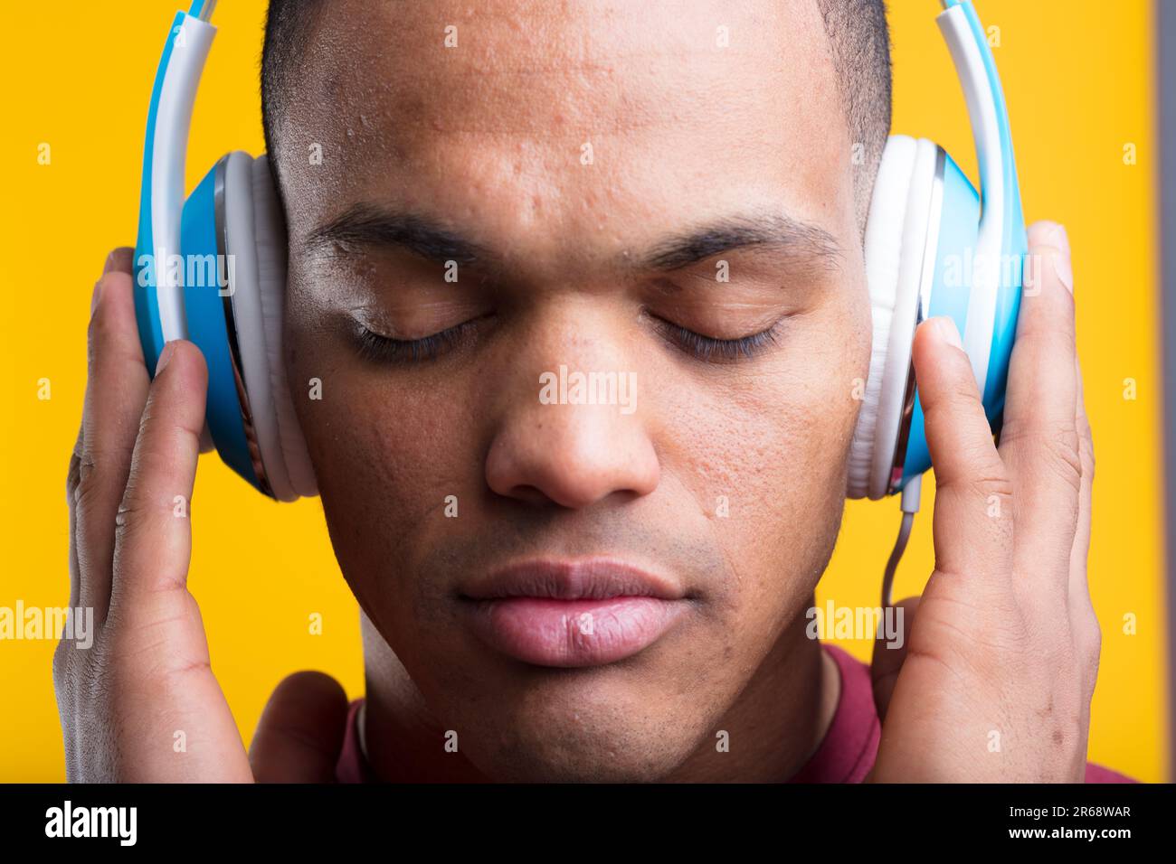 Mann mit geschlossenen Augen, genießt Musik oder Podcast, mit blauen Kopfhörern auf gelbem Hintergrund. Er hat kurze rasierte Haare und dunkelbraune Haut. Sein T-sh Stockfoto