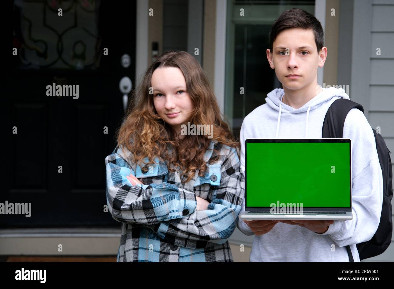 Ernsthafte Teenager stehen mit Laptop und Rucksack Schultasche zeigen grünen Bildschirm Chrom Schlüssel auf Laptop Herbst Frühlingsstudium Stand in der Nähe von zu Hause Werbung Online-Sprachkurse Junge und Mädchen Stockfoto