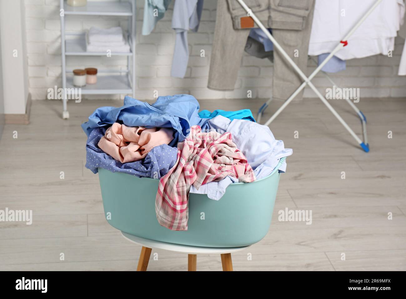 Wäschekorb aus Plastik, überfüllt mit Kleidung auf weißem Hocker im Innenbereich Stockfoto