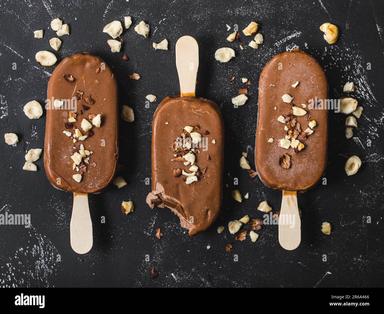 Milchschokolade Eis am Stiel mit Haselnüssen. Nahaufnahme. Eis am Stiel mit Schokolade, Stäbchen und schwarzem Steinhintergrund. Draufsicht. Schokolade Stockfoto