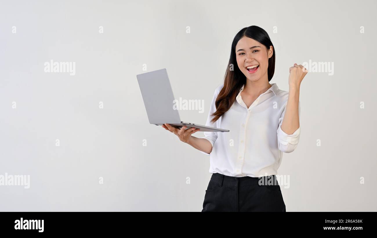 Eine fröhliche junge Asiatin in einem weißen Hemd steht vor einem isolierten weißen Hintergrund und zeigt ihre Faust, während sie ihren Laptop hält. Erfolg, Feier Stockfoto
