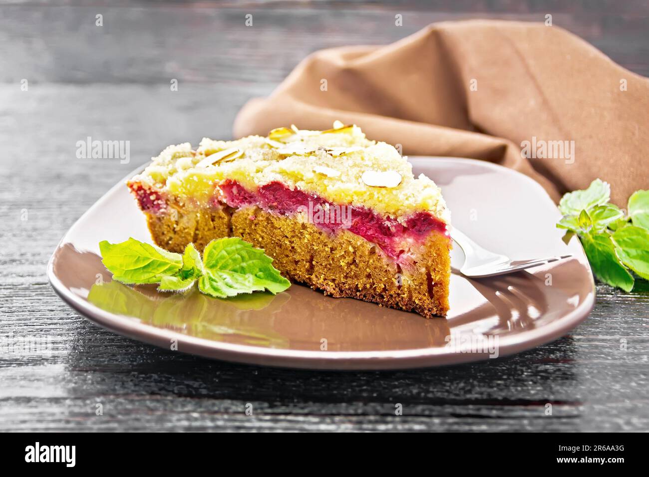 Ein Stück süßen Kuchen mit Himbeeren, Butterbröckeln und Mandelflocken auf einem Teller, Minze und Serviette auf Holzbretthintergrund Stockfoto