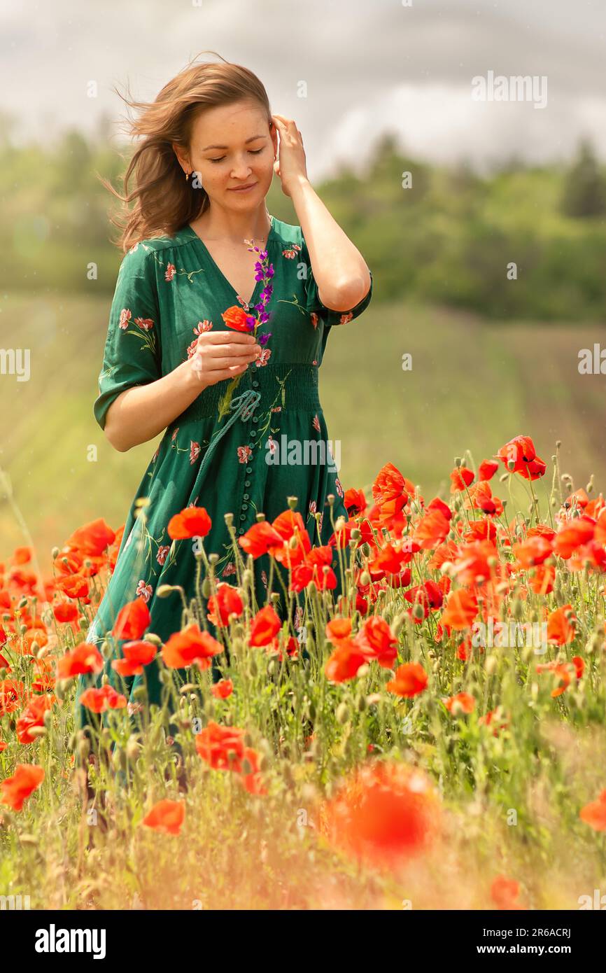 Eine junge weiße Frau auf einem Mohnfeld, mit einer Hand glättet sie ihr Haar, in der anderen hält sie eine rote Blume mit einem Lächeln, eine Atmosphäre der Freude, der Liebe Stockfoto