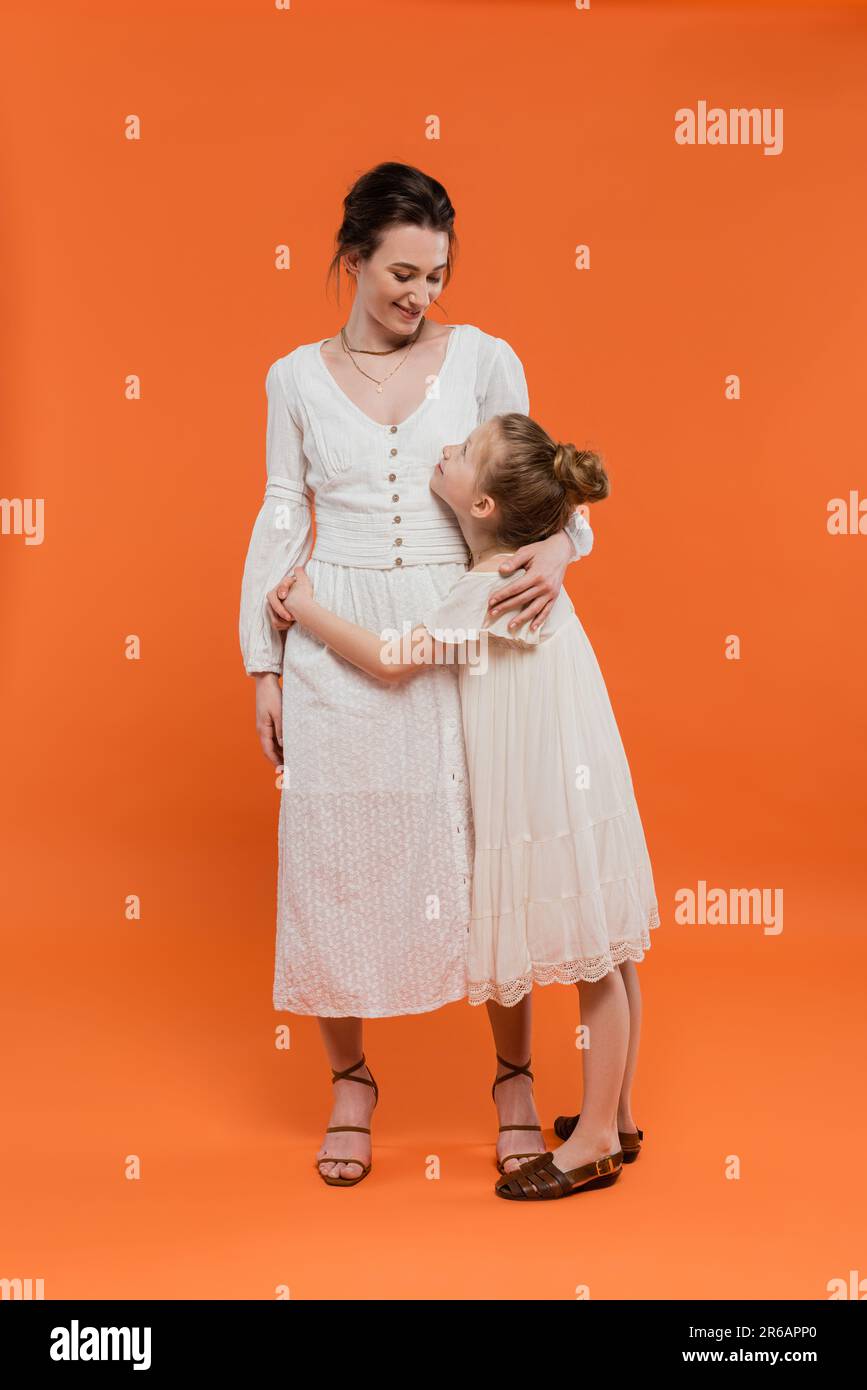 Weibliche Bindung, fröhliches junges Mädchen umarmt Mutter auf orangefarbenem Hintergrund, volle Länge, Glück, weiße Sonnenkleider, Sommermode, Zusammengehörigkeit, Stockfoto