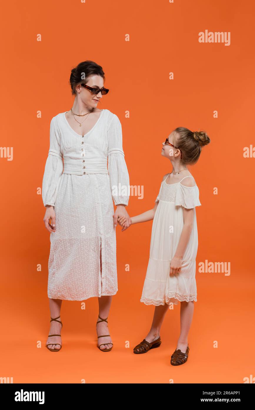 Mütterliche Liebe, stilvolle Familie, Mutter und Kind in Sonnenbrille halten Hände auf orangefarbenem Hintergrund, weiße Sonnenkleider weibliche Bindung, modisch, einfach Stockfoto